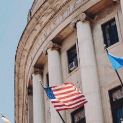 关闭-up view of a neoclassical government building with large columns and a facade made of stone. 几个旗帜, including the American flag, are visible in front of the building against a clear blue sky.